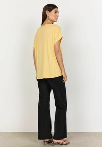 Gul, løstsittende t-shirt med korte �ærmer og en afslappet halsudskæring, kombineret med sorte, vide bukser og mønstrede sandaler med åben tå.