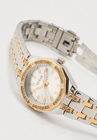 Sølv- og guldfarvet ur med oktagonalt hvidt urværk, guldmarkører og diamantdetaljer på rammen. Rustfrit stål og guldbetonet armbånd.