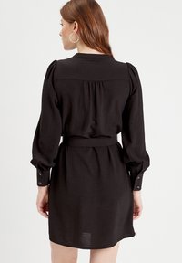 Robe noire avec manches bouffantes, taille ceinturée et poignet à boutons. Le tissu semble texturé, avec une coupe fluide tombant au-dessus des genoux.