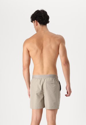 Des shorts de bain gris clair présentent une texture tissée, une taille élastique avec logo et une poche arrière, conçus pour le confort et la mobilité.