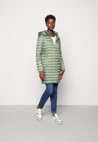 Hellgrüne Steppjacke mit Kapuze, ausgestattet mit Zickzacknähten und zwei seitlichen Reißverschlusstaschen. Kombiniert mit blauen Jeans und Sneakers mit grünen Akzenten.