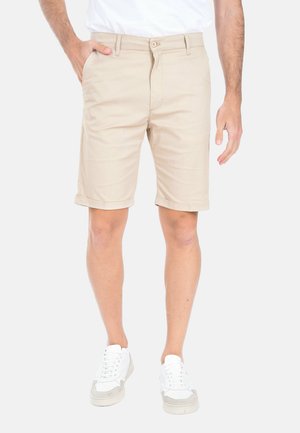 Uomo che indossa pantaloncini beige con orlo arrotolato e tasche frontali, una t-shirt bianca e scarpe da ginnastica bianche, in piedi su sfondo bianco.