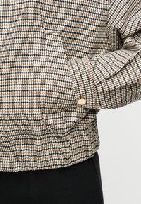 Πλεκτό σακάκι με μοτίβο pied-de-poule σε εκρού, μαύρο και πορτοκαλί. Διαθέτει μανσέτα με κουμπιά και ελαστική μέση για ένα εφαρμοστό look.