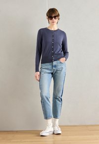 Merino paita tummansinisestä neuleesta, jossa pyöreä kaulus ja napitus, yhdistettynä vaaleansinisiin farkkuihin, joissa on tummat sivusuorat raidat, sekä valkoiset tennarit.