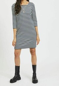 Pruhované šaty s třičtvrtinovými rukávy, s námořnickými a bílými horizontálními pruhy. Hladká tkanina, do kolen, kombinované s černými kotníkovými botami.