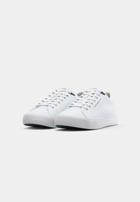 Paire de baskets basses blanches avec lacets blancs et marquage discret sur le côté et la languette, sur un fond clair uni.