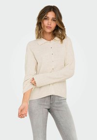 Hellbeige, gerippte Knopfleiste mit Kragen, langen Ärmeln und einem leicht abgerundeten Saum. Kombiniert mit grauen, eng anliegenden Jeans.