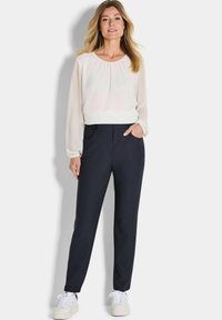 Leichte, fließende Cremebluse mit langen Ärmeln und gerafftem Ausschnitt, kombiniert mit dunkelblauen, maßgeschneiderten Hosen und weißen Sneakers.