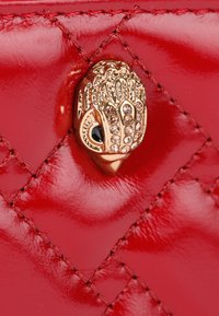 Accessorio in pelle rossa trapuntata con una chiusura decorativa dorata caratterizzata da dettagli intricati e rhinestones incastonati. Texture complessivamente liscia.