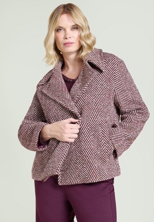 Kvinna med blont hår som bär en vinröd och vit fiskbensmönstrad kappa över en vinröd outfit, stående mot en enfärgad bakgrund.