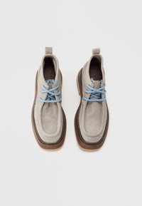 Bottes mi-mollet en feutre gris clair avec des lacets bleus, des semelles en caoutchouc et des accents marron. Dotées d'un bout rond et de coutures renforcées le long des bords.