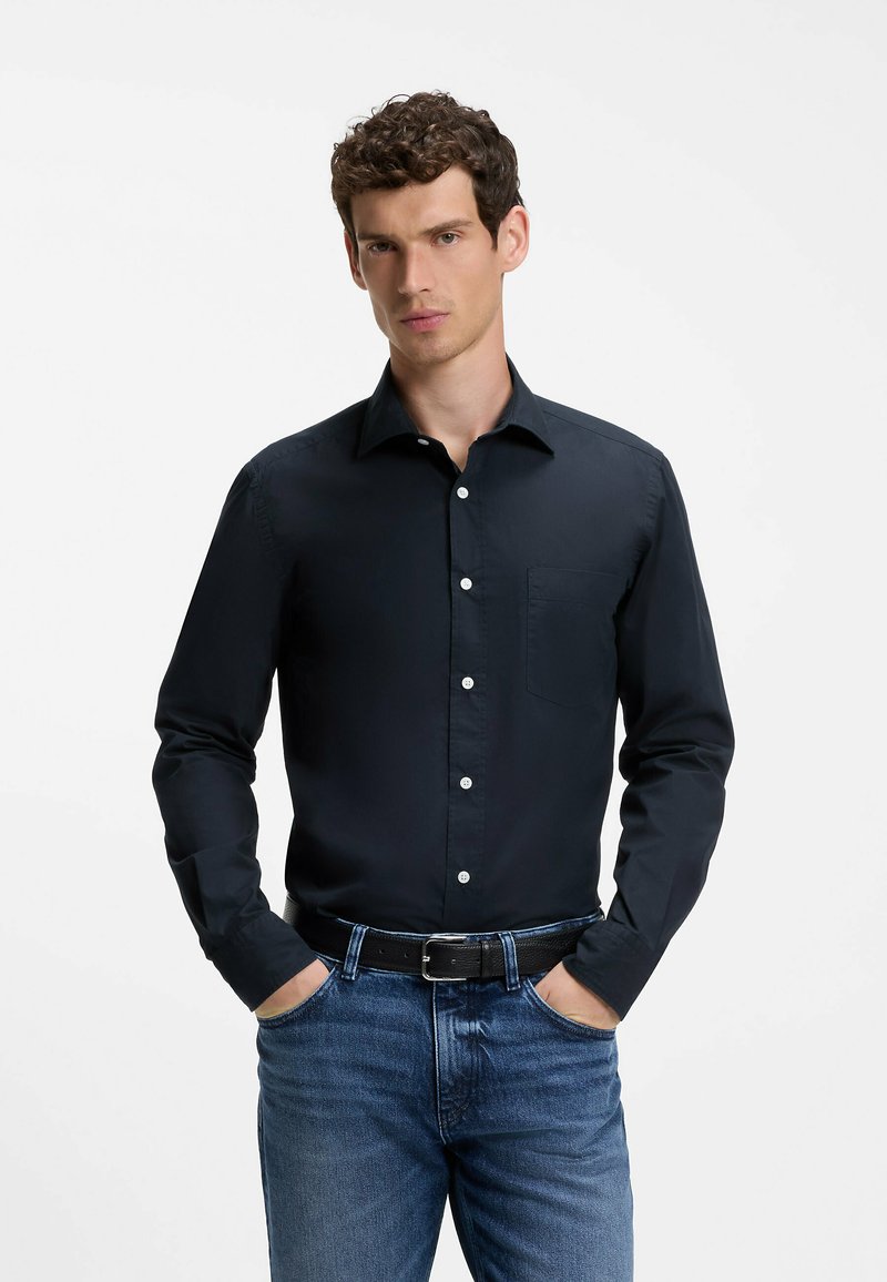 Uomo che indossa una camicia nera con bottoni e taschino sul petto, jeans blu e cintura nera, in piedi con le mani nelle tasche davanti a uno sfondo bianco.