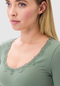 Top verde a costine con scollatura in pizzo ondulato. Il tessuto appare testurizzato e il design è aderente con un taglio a forma di V.