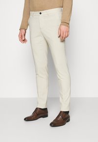 Beige, slim fit szabású nadrág, lapos elülső résszel, oldal zsebekkel és gombos záródással, sötétbarna elegáns cipővel párosítva.