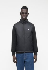 Schwarze Steppweste mit hohem Kragen, Reißverschluss und schwarzen langen Ärmeln. Verfügt über ein dezentes Logo und eine glatte Textur, kombiniert mit blauer Jeans.