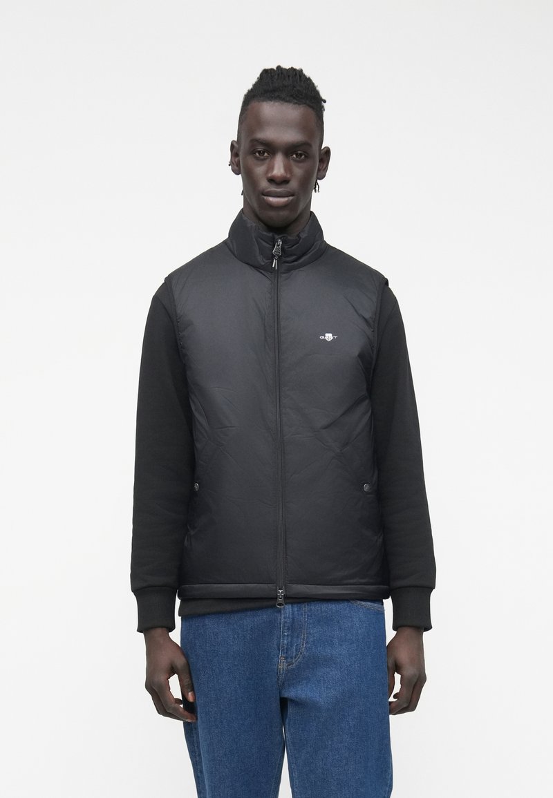 Schwarze Steppweste mit hohem Kragen, Reißverschluss und schwarzen langen Ärmeln. Verfügt über ein dezentes Logo und eine glatte Textur, kombiniert mit blauer Jeans.