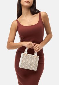 Hvid håndtaske med perleudsmykning, rektangulær form og korte håndtag. Båret af en model iført en brun ribstrikket kjole.