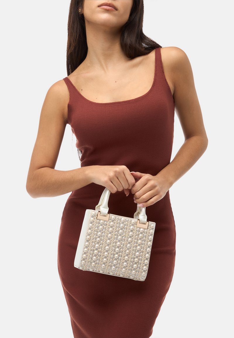 Hvid håndtaske med perleudsmykning, rektangulær form og korte håndtag. Båret af en model iført en brun ribstrikket kjole.