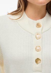 Nahaufnahme eines cremefarbenen Strickpullovers mit geripptem Kragen und vier goldenen muschelförmigen Knöpfen auf der Vorderseite.