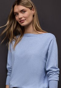 Jeune femme aux longs cheveux blonds portant un pull bleu clair, debout devant un fond gris foncé, souriant légèrement.