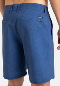 Modré kraťasy vyrobené z textúrovanej látky s jemným vzorom. Sú vybavené dvomi zadnými vreckami a logom na páse.