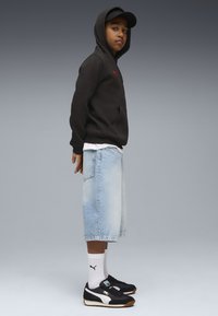 Felpa con cappuccio nera e logo rosso, pantaloni corti in denim azzurro chiaro, sneakers bianco e nere, e calze bianche alla caviglia. Posizione in piedi.