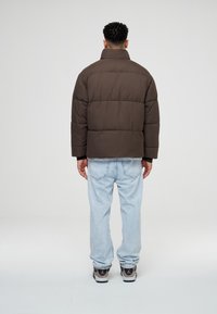 Brun puffjacka med hög krage och kviltad design, matchad med ljusblå jeans och svartvita sneakers.