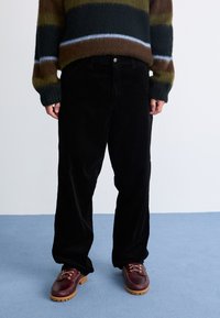 Sorte fløjl brede bukser med en struktureret overflade, parret med burgunder snøresko og en stribet, fuzzy brun sweater.