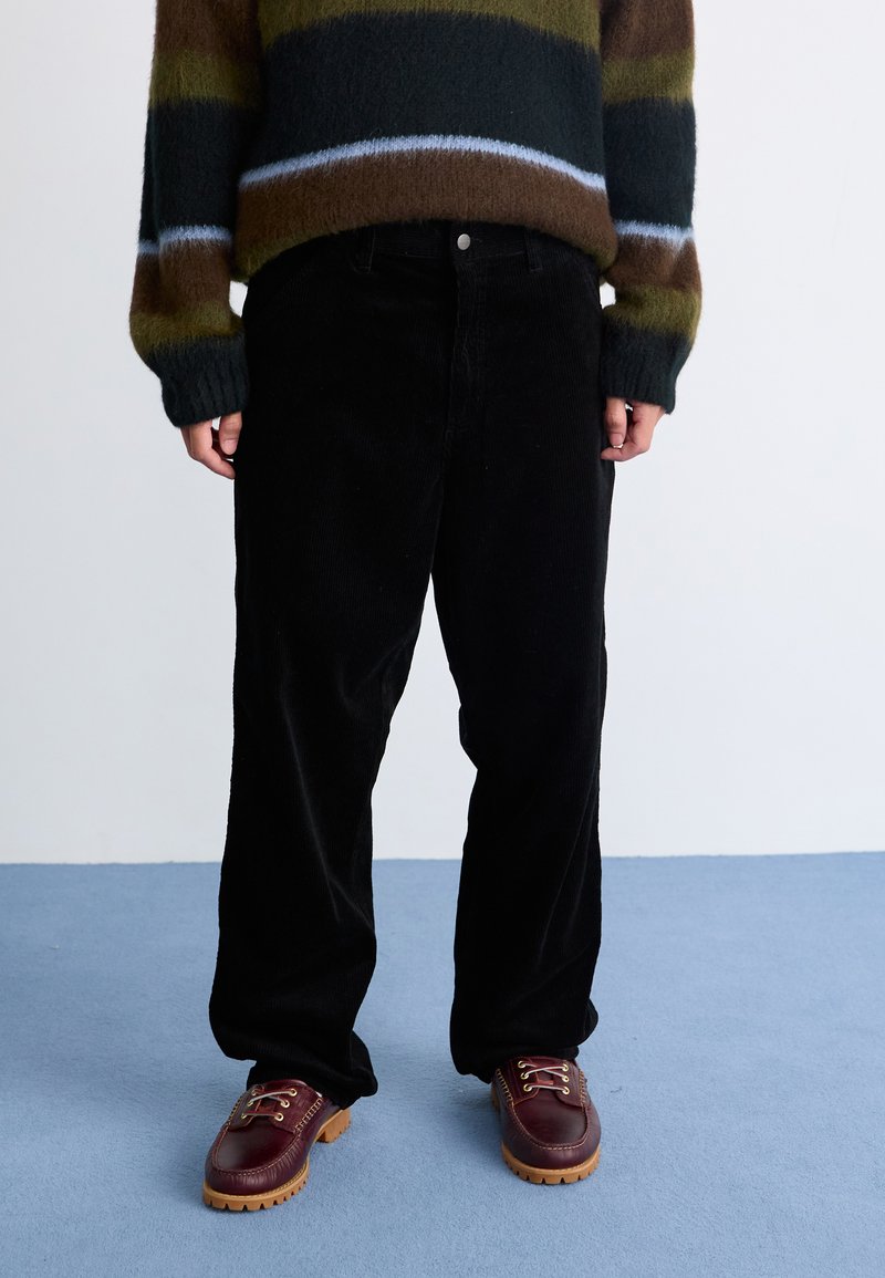 Sorte fløjl brede bukser med en struktureret overflade, parret med burgunder snøresko og en stribet, fuzzy brun sweater.