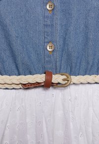 Jeansowa bluzka z srebrnymi guzikami, kremowy pleciony pasek z brązowymi akcentami oraz biała, teksturowana spódnica w kwiatowy wzór.