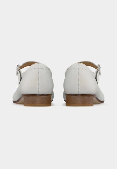 Ζευγάρι λευκά δερμάτινα παπούτσια Mary Jane με χαμηλά ξύλινα τακούνια και ασημένιες, αγκράφες με λουριά, φαίνεται από πίσω σε απλό φόντο.