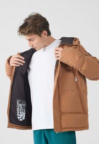 Giacca imbottita marrone con chiusura a zip, rivestimento interno nero e dettaglio grafico sulla tasca interna. Materiale morbido e isolante.