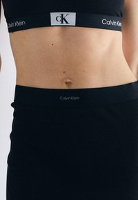 Sutien sport negru Calvin Klein, cu o bandă elastică lată ce prezintă numele și logo-ul brandului în alb. Textură ribbed și design fitted.