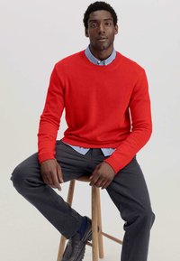 Roter Strickpullover mit Rundhalsausschnitt, gerippte Bündchen und Saum, über einem hellblauen Hemd mit Kragen getragen. Graue Hose und schwarze Schuhe.