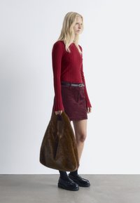 Rotes geripptes Langarmoberteil, bordeauxfarbene Cordrock, schwarzer Gürtel. Hält eine braune Kunstfell-Tasche, trägt schwarze Slipper und Socken.