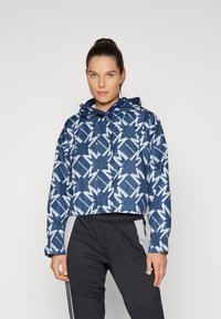 Zkrácená modrá bunda s geometrickým bílým vzorem, s kapucí, zapínáním na zip a elastickými manžetami. V kombinaci s černými sportovními kalhotami.