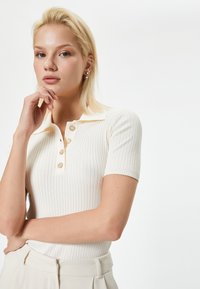 Geribbeld, crème top met korte mouwen en een kraaghalslijn, voorzien van vijf knoopaccenten. Zachte textuur, aansluitend ontwerp en subtiele off-white tint.