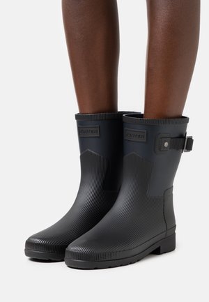 Stivali da pioggia neri in gomma a metà polpaccio con texture a coste e fibbia laterale, indossati su gambe dalla pelle scura su sfondo bianco.