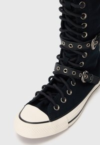 Schwarzer High-Top-Sneaker mit weißer Gummispitze, schwarzen Schnürsenkeln und zwei schwarzen Riemen mit silbernen Ösen und Schnallen.