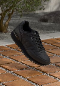 Zapatilla atlética negra con un upper texturizado, con un acabado de cuero liso y cordones negros, sobre un fondo de losas de madera y grava.