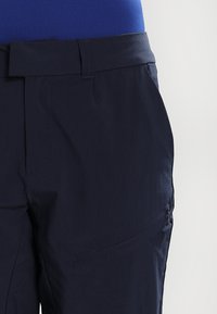 Laivansininen kevyet ulkoiluhousut, joilla on sileä pinta, vyölenkit ja sivutasku vetoketjulla. Alhaalla on brändin logo.