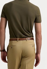 Mann in olivgrünem Poloshirt und beigen Hosen mit braunem Ledergürtel, von hinten vor einem einfarbigen Hintergrund dargestellt.