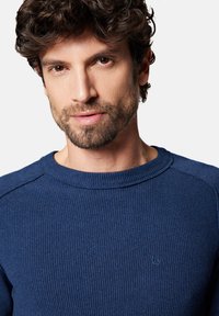 Niebieski sweter z okrągłym dekoltem o prążkowanej fakturze i subtelnym detalem logo na piersi. Materiał wydaje się przytulny i lekki, idealny do codziennego noszenia.