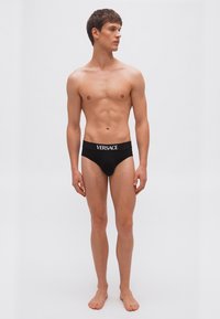 Jeune homme debout, pieds nus, portant un slip noir Versace, regardant vers sa gauche sur un fond gris clair uni.