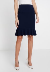 Marinblå knä-lång kjol i stretchigt tyg, med en utsvängd volang i fållkanten. Matchas med vita högklackade skor.