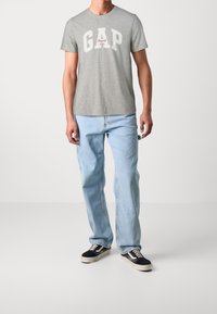 T-shirt en coton gris avec le logo "GAP" en blanc et rouge, assortie à un jean en denim bleu clair et des baskets noires avec des semelles blanches.