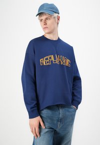 Ung mann som har på seg en blå hettegenser med teksten "REPLAYONE", lys blå caps og blå jeans, ser til siden med en hånd i lommen.