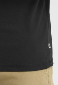 Zwarte katoenen shirtzoom met klein wit merklabel boven beige broek, waarbij de textuur van de stof en de stiksels zichtbaar zijn.
