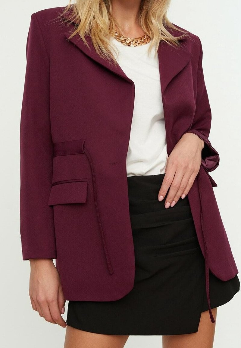 Veste bordeaux au design structuré, avec une poche unique et un col cranté. Portée sur une chemise blanche et une jupe noire, avec un tissu doux.