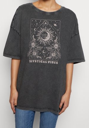 Person wearing a faded black oversized t-shirt with sun, moon, stars, flowers, and "Mystical Vibes" text in light print on front.